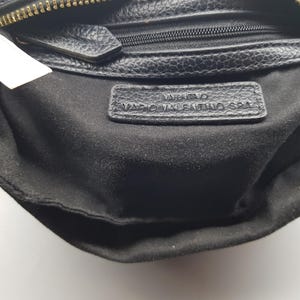 Pu&ograve; includere: Primo piano di una borsa in pelle nera con cerniera dorata. L'interno &egrave; in tessuto nero. Una piccola etichetta in pelle con le parole "Valentino Mario Valentino SPA" &egrave; visibile all'interno della borsa.