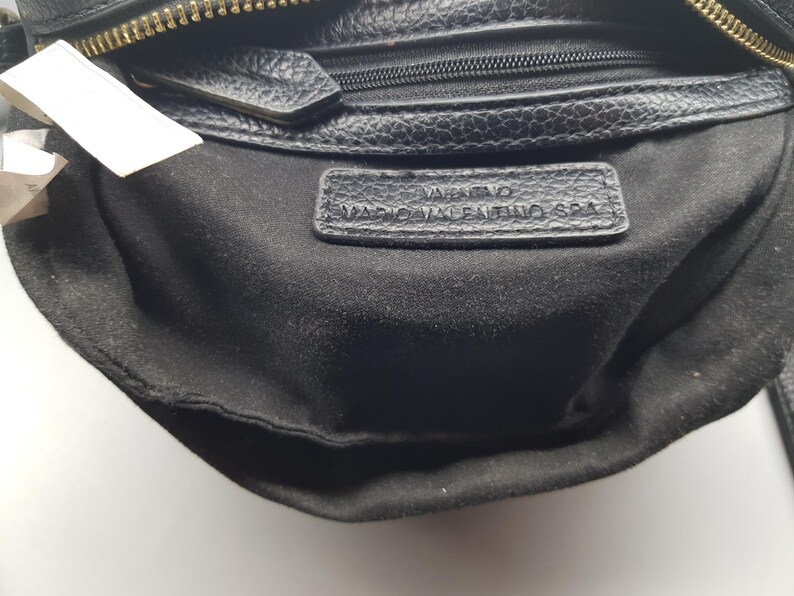 Pu&ograve; includere: Primo piano di una borsa in pelle nera con cerniera dorata. L'interno &egrave; in tessuto nero. Una piccola etichetta in pelle con le parole "Valentino Mario Valentino SPA" &egrave; visibile all'interno della borsa.