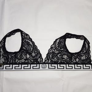 May include: Black lace bralette with floral patterns. The bralette features a white band with a Greek key design and a Medusa head emblem. The straps are black.