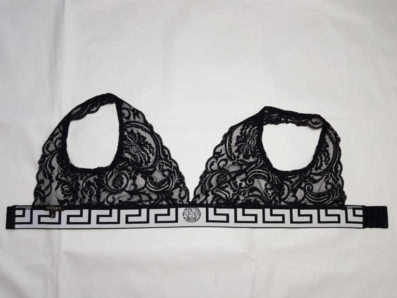 May include: Black lace bralette with floral patterns. The bralette features a white band with a Greek key design and a Medusa head emblem. The straps are black.