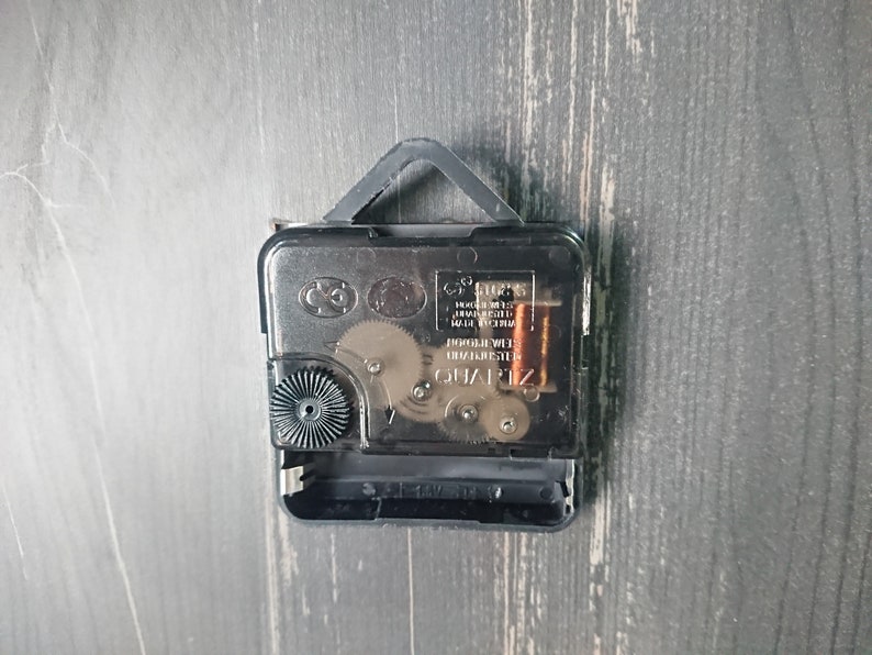 K&ouml;nnte beinhalten: Nahaufnahme der R&uuml;ckseite eines Uhrwerks mit einem schwarzen Kunststoffgeh&auml;use und einem silbernen Metallgetriebe. Die Uhr ist mit "QUARTZ" und "MADE IN CHINA" beschriftet.