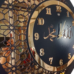 K&ouml;nnte beinhalten: Eine schwarze und goldene Uhr mit arabischen Ziffern und dem Wort "Allah" in arabischer Schrift. Die Uhr hat ein dekoratives, geometrisches Design am Rand.