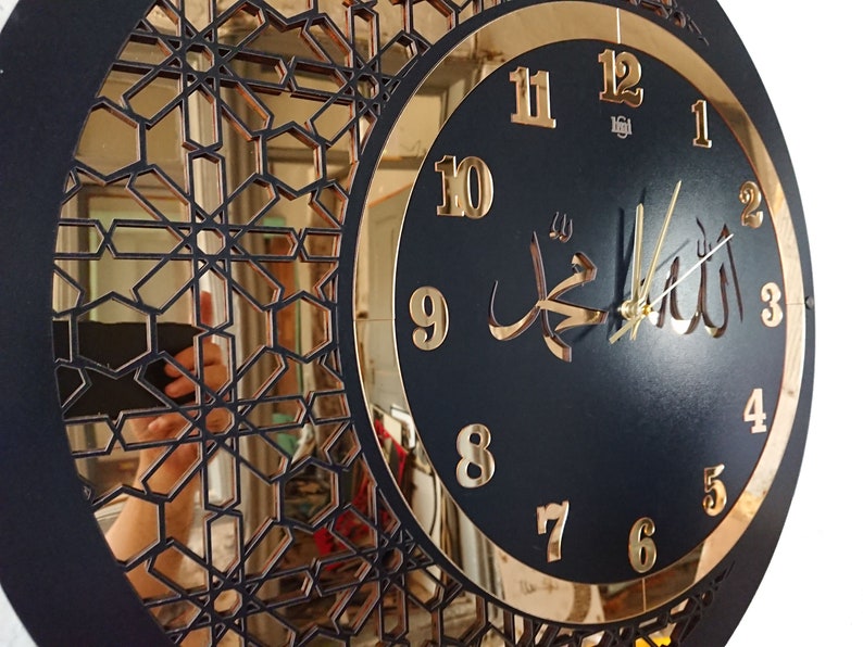 K&ouml;nnte beinhalten: Eine schwarze und goldene Uhr mit arabischen Ziffern und dem Wort "Allah" in arabischer Schrift. Die Uhr hat ein dekoratives, geometrisches Design am Rand.