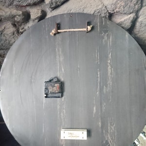 K&ouml;nnte beinhalten: Die R&uuml;ckseite einer runden Holzuhr mit schwarzer Oberfl&auml;che. Die Uhr hat ein schlichtes Design mit einem Seil, das an zwei Metallhaken h&auml;ngt. Der Uhrmechanismus ist auf der R&uuml;ckseite sichtbar.