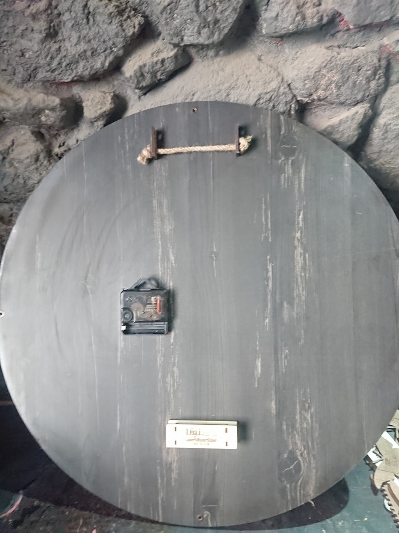 K&ouml;nnte beinhalten: Die R&uuml;ckseite einer runden Holzuhr mit schwarzer Oberfl&auml;che. Die Uhr hat ein schlichtes Design mit einem Seil, das an zwei Metallhaken h&auml;ngt. Der Uhrmechanismus ist auf der R&uuml;ckseite sichtbar.