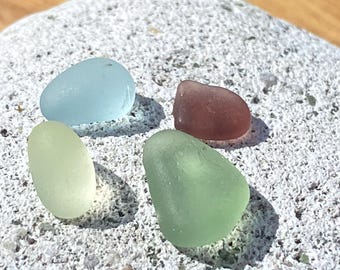 Könnte beinhalten: Vier glatte, vom Meer abgenutzte Glasstücke in Blau-, Grün- und Brauntönen. Die Stücke sind oval geformt und liegen auf einer strukturierten, hellfarbigen Oberfläche. Die Farben sind weich und gedämpft.