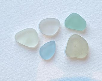 Könnte beinhalten: Fünf Stücke Meeresglas in Pastellfarben: Hellblau, Hellgrün und Weiß. Die glatten, abgerundeten Formen sind auf einer strukturierten weißen Oberfläche verstreut und erzeugen eine ruhige, natürliche Ästhetik.