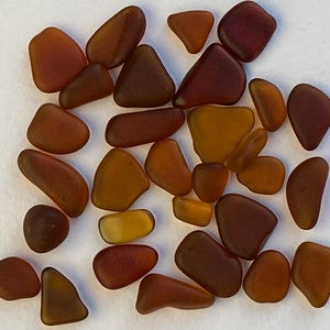 May include: A collection of numerous small, smooth, frosted glass pieces in various shades of brown, amber, and deep red. Each piece features rounded edges and a matte, weathered texture, scattered across the frame in an irregular arrangement.