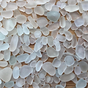 Puede incluir: Primer plano de una colección de piezas de vidrio marino lisas y esmeriladas. Las piezas, de diversas formas y tamaños, son principalmente blancas, azules y grises, dispuestas sobre una superficie de madera marrón claro. Las formas varían de pequeños guijarros a formas más alargadas.