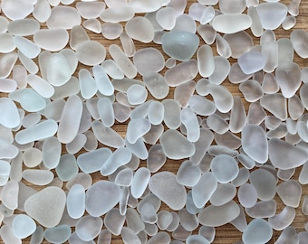 May include: A collection of smooth, frosted sea glass pieces in various shapes and sizes. The sea glass is primarily shades of white, blue, and gray, scattered on a light brown wooden surface. The pieces vary from small pebbles to larger, elongated forms.