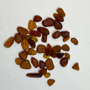 May include: A collection of small, smooth, brown and amber sea glass pieces. The sea glass is scattered on a white surface.