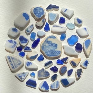 May include: A collection of smooth, weathered sea glass pieces arranged in a circular pattern. The assortment features various shades of blue and white, including solid cobalt, light blue, and white fragments with blue patterns and ceramic designs.