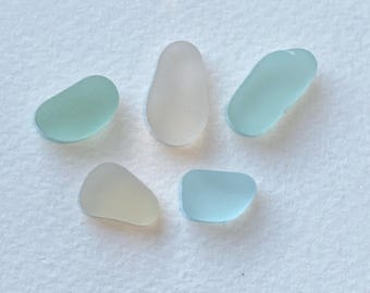 Könnte beinhalten: Fünf Stücke Meerglas in verschiedenen Formen und Pastellfarben. Die Farben umfassen Hellblau, Hellgrün und Off-White. Das Meerglas ist glatt und abgerundet, was darauf hindeutet, dass es vom Ozean geschliffen wurde.