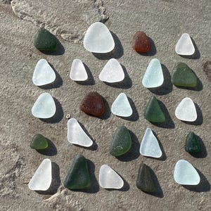 May include: A collection of sea glass pieces in assorted shapes and colours, including clear, green, and brown. The pieces are arranged on a textured, grey surface, creating a natural, beachy aesthetic.