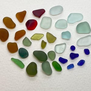 May include: A selection of sea glass pieces in a range of colours, such as brown, red, green, blue, and clear. The smooth, rounded glass shapes are arranged on a white surface, forming a colourful display of natural beach finds.