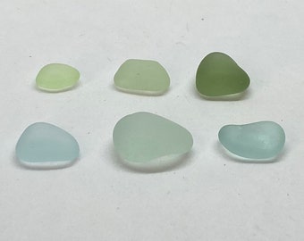 Könnte beinhalten: Sechs Stücke Meeresglas in verschiedenen Grün- und Blautönen sind auf einer weißen Oberfläche angeordnet. Die Stücke sind glatt und abgerundet, mit einem mattierten Aussehen. Die Farben reichen von blassgelb-grün bis zu tiefem Olivgrün und Hellblau.
