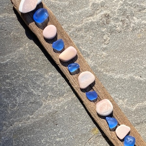 Puede incluir: Un trozo de madera a la deriva con una fila de vidrio de mar y conchas marinas. El vidrio de mar es azul y las conchas son rosadas y blancas.
