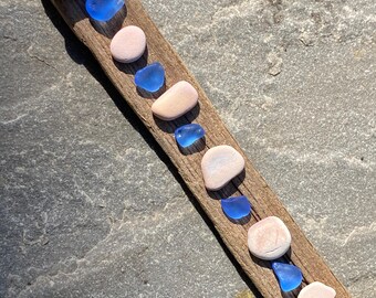 Könnte beinhalten: Ein Stück Treibholz mit einer Reihe von Meerglas und Muscheln. Das Meerglas ist blau und die Muscheln sind rosa und weiß.