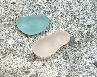 May include: Two sea glass pieces, one is a light blue, the other is a light pink.