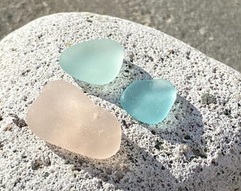 May include: Three pieces of sea glass in shades of light blue and pink rest on a textured, light gray rock. The sea glass has smooth, rounded edges, and the colors are soft and translucent.