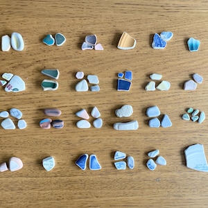 May include: A collection of assorted sea glass and ceramic shards arranged in rows. The pieces feature various shapes, sizes, and colors including white, blue, green, yellow, and pink, with some fragments displaying floral patterns and textured surfaces.