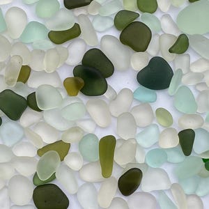 May include: A collection of sea glass in various shades of green, white, and blue. The glass pieces are smooth and rounded, suggesting they have been tumbled by the ocean for a long time.