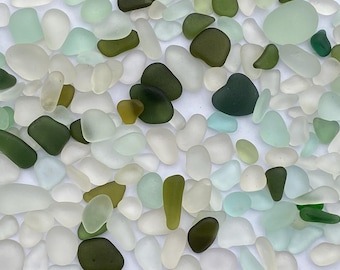 Könnte beinhalten: Eine Sammlung von Meerglas in verschiedenen Grüntönen, Weiß und Blau. Die Glassplitter sind glatt und abgerundet, was darauf hindeutet, dass sie lange Zeit vom Meer geschliffen wurden.