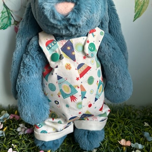 Op de afbeelding: Een blauw pluche konijn met een witte overall met een patroon van raketten, planeten en sterren. De overall heeft groene knopen en witte randen. Het konijn staat op een bed van groen gras met kleurrijke bloemen.