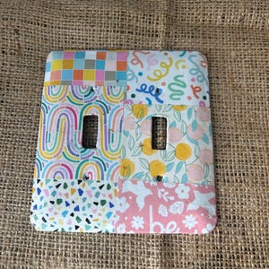 May include: A white light switch cover with a patchwork design featuring colorful patterns, including rainbows, geometric shapes, and floral motifs. The cover has two switches.