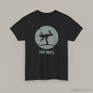 May include: A black short-sleeve t-shirt featuring a circular graphic in a muted sage green color. The graphic depicts a silhouette of a person balancing on a line, with the text TOM WAITS printed in a distressed font directly below the circle.