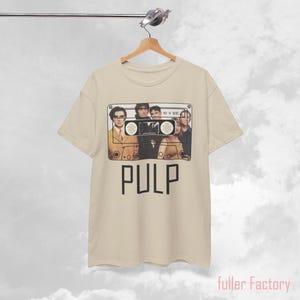 May include: A beige short-sleeve t-shirt featuring a graphic of a cassette tape with a band portrait inside the frame. The text 'HIS 'N' HERS' is printed on the cassette, and the word 'PULP' is printed in large black letters below the graphic.