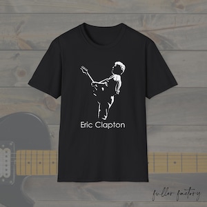 Puede incluir: Camiseta negra con una silueta blanca de Eric Clapton tocando la guitarra. El texto "Eric Clapton" está impreso en blanco. La camiseta se muestra sobre un fondo de madera.