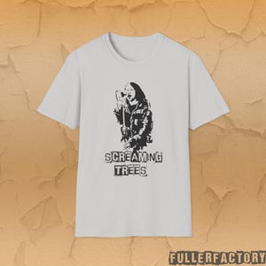 Puede incluir: Camiseta blanca con un gráfico en blanco y negro de una persona cantando en un micrófono. El texto "SCREAMING TREES" está impreso debajo en una fuente de bloque. Cuello redondo.