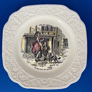 Puede incluir: Un plato de cerámica blanco con un borde floral en relieve. El centro del plato presenta una ilustración colorida de una mujer y un niño vendiendo cerillas en una calle de Londres. El texto "CRIES OF LONDON" y "DO YOU WANT ANY MATCHES" está impreso en el plato.