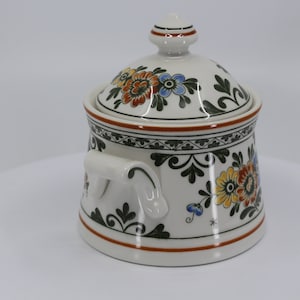 Könnte beinhalten: Weiße Zuckerdose aus Keramik mit Deckel und zwei Griffen. Die Schale ist mit einem floralen Muster in Orange-, Gelb-, Grün- und Blautönen verziert.