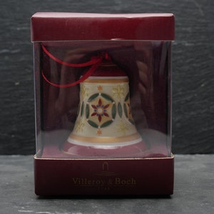 Op de afbeelding: Een witte porseleinen bel-versiering met een geometrisch ontwerp in rood, groen en goud. De bel zit in een doorzichtige plastic doos met een rood deksel en de woorden "Villeroy & Boch" gedrukt op de bodem.