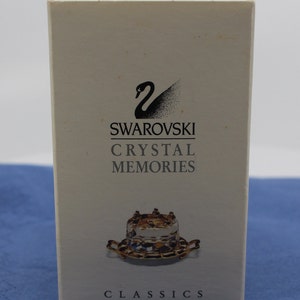 Puede incluir: Una caja blanca con el logotipo de Swarovski y el texto "Crystal Memories Classics". La caja presenta un pequeño pastel de cristal transparente con detalles dorados.