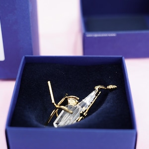 Peut inclure: Une figurine de saxophone en cristal transparent et doré repose dans une boîte bleu foncé. La boîte est dans une boîte bleue plus grande, sur fond rose. Le saxophone présente un design détaillé.