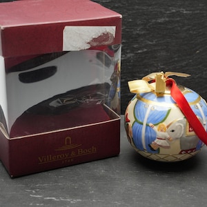 Könnte beinhalten: Ein weißer Porzellan-Weihnachtsbaumschmuck mit blauem und goldenem Design. Der Schmuck zeigt eine Szene mit einem weißen Elefanten, einer Frau in einem blauen Kleid und einem Kind in einem weißen Kleid. Der Schmuck ist in einer roten Schachtel mit einem durchsichtigen Plastikfenster verpackt. Die Schachtel trägt den Schriftzug "Villeroy & Boch".