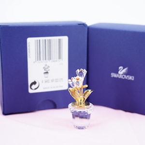 Op de afbeelding: Een kristallen bloemenfiguur in een doorzichtige pot met goudkleurige bladeren en stengel, geplaatst voor een blauwe doos. De doos heeft een wit label met een barcode en het Swarovski-logo. De figuur is ongeveer 5 cm hoog.