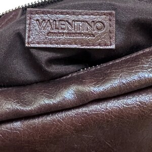Pu&ograve; includere: Una borsa in pelle marrone con un'etichetta logo Valentino. La borsa ha una chiusura a zip e una superficie liscia e testurizzata.