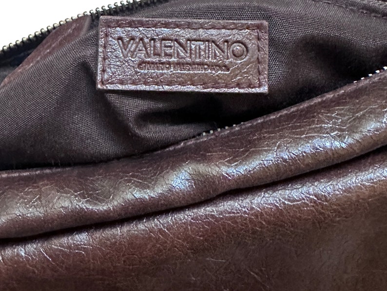 Pu&ograve; includere: Una borsa in pelle marrone con un'etichetta logo Valentino. La borsa ha una chiusura a zip e una superficie liscia e testurizzata.