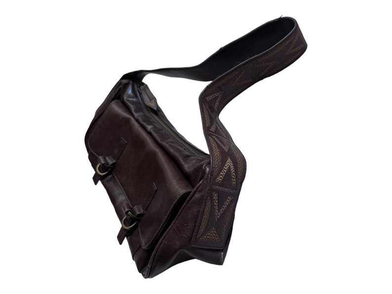 Pu&ograve; includere: Una borsa a tracolla in pelle marrone con una tracolla decorativa con un motivo geometrico. La borsa ha due fibbie sul davanti.