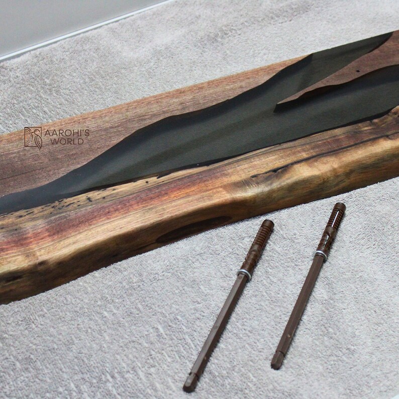 May include: A long, wooden board with a dark green epoxy river running through it. The board has a natural wood grain and a reddish-brown colour. Two brown metal screws are lying on a grey surface below the board.
