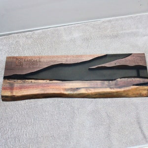 May include: A long, rectangular piece of wood with a dark brown, black and grey epoxy resin inlay. The wood has a natural, live edge and the resin is shaped like a river. The piece is signed "Aarohi's World" in the top left corner.