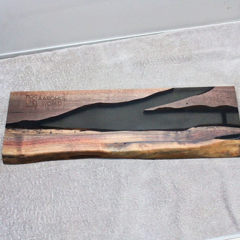 May include: A long, rectangular piece of wood with a dark brown, black and grey epoxy resin inlay. The wood has a natural, live edge and the resin is shaped like a river. The piece is signed "Aarohi's World" in the top left corner.