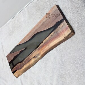 May include: A long, wooden serving tray with a natural edge and a black epoxy resin inlay. The tray is approximately 61 cm long and 15 cm wide. The wood has a rich brown colour and the resin is smooth and glossy.