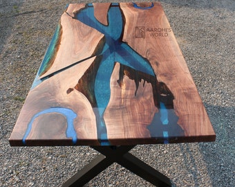 May include: A rectangular dining table featuring a natural live-edge wood slab with a translucent blue epoxy resin river inlay. The table is supported by a black metal X-shaped base and includes the text AAROHI'S WORLD printed on the surface.
