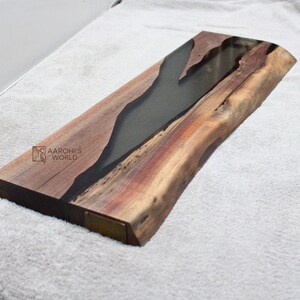 May include: A long, rectangular wooden serving tray with a natural wood grain finish. The tray has a black epoxy resin inlay that resembles a river. The tray has a brass plaque on the side with the text "AAROHI'S WORLD".