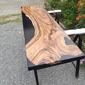 May include: A rectangular table featuring a natural live-edge wood slab with prominent brown grain patterns integrated into a smooth, solid black resin surface. The table is supported by a black metal frame with four legs and white protective padding.